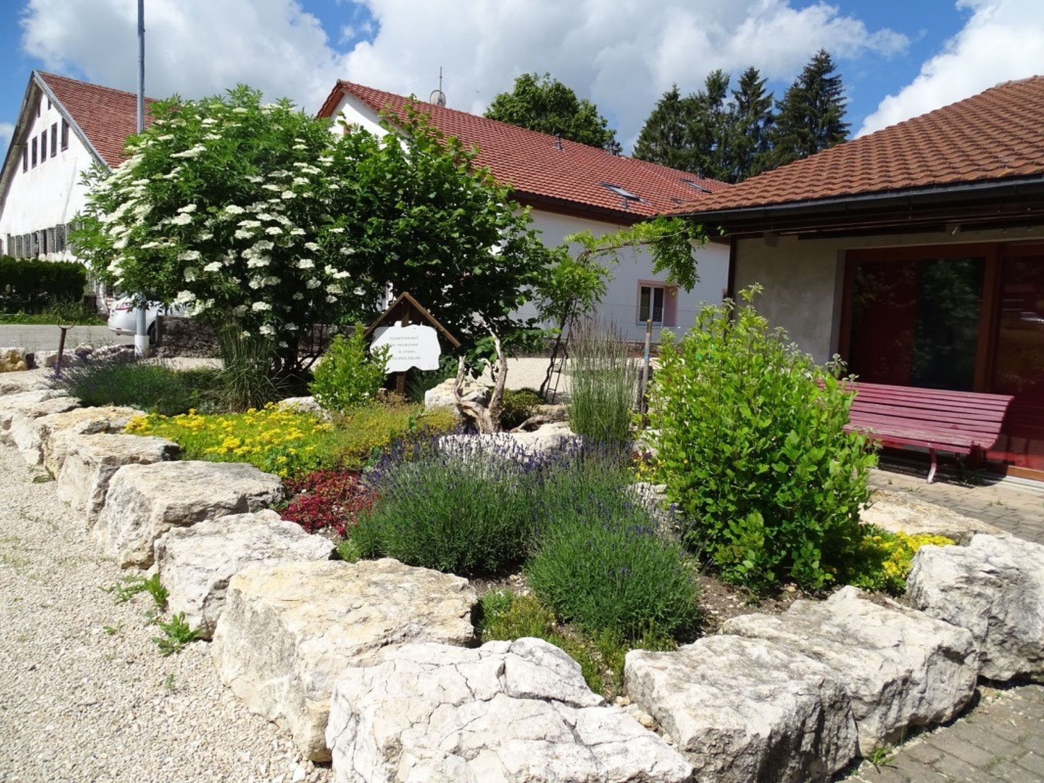 A building with a red-tiled roof, surrounded by a garden with various plants and a stone border.