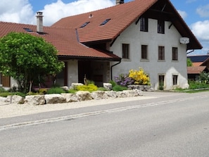 A two-story building with a red-tiled roof, a stone foundation, and a garden with various plants. - Appartement - La Racrue, (Lajoux JU), by Interhome (Lajoux JU)