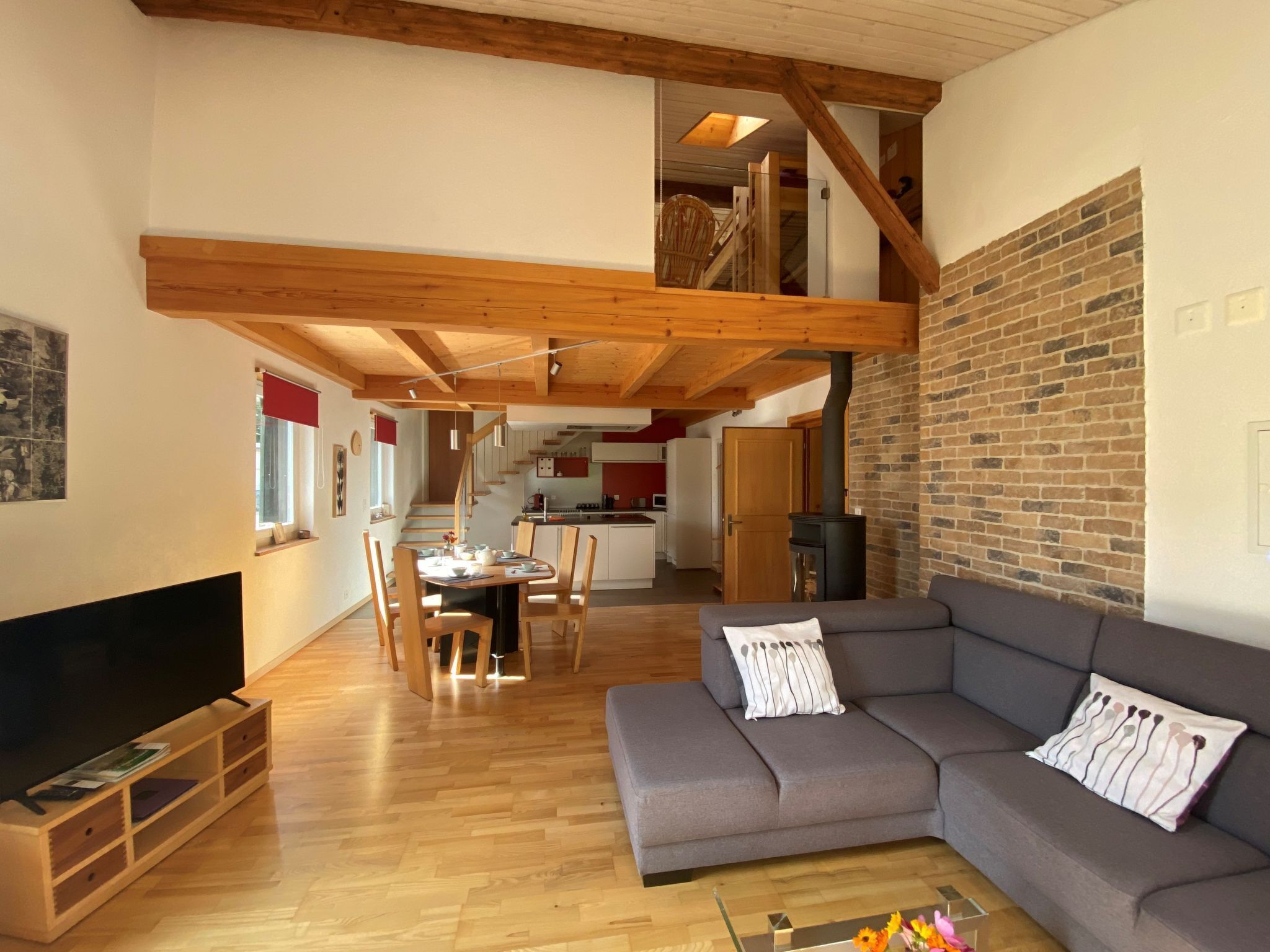 Ein großzügiger Wohnbereich mit grauem Sofa, Holzboden und einer Küche im Hintergrund.