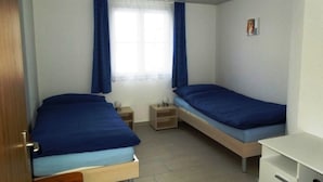 Een kamer met twee eenpersoonsbedden, een klein nachtkastje met een lamp, een wit bureau en een raam met gordijnen.