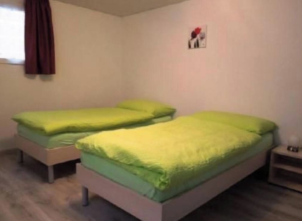 A room with two single beds, each with a green bedspread, a small bedside table, and a wall-mounted light.