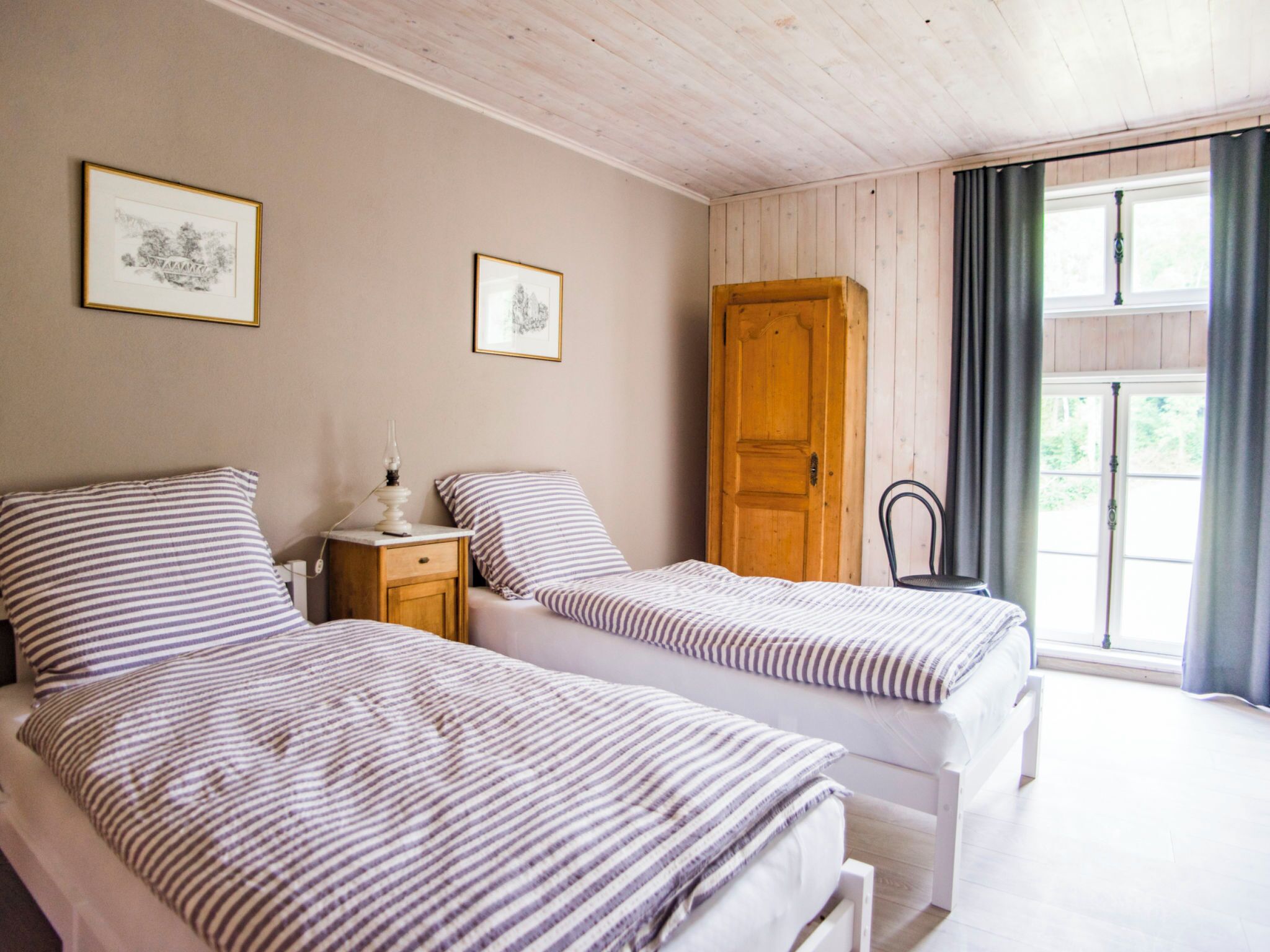 A bedroom with two beds, a wooden wardrobe, a bedside table with a lamp, and a window with curtains.