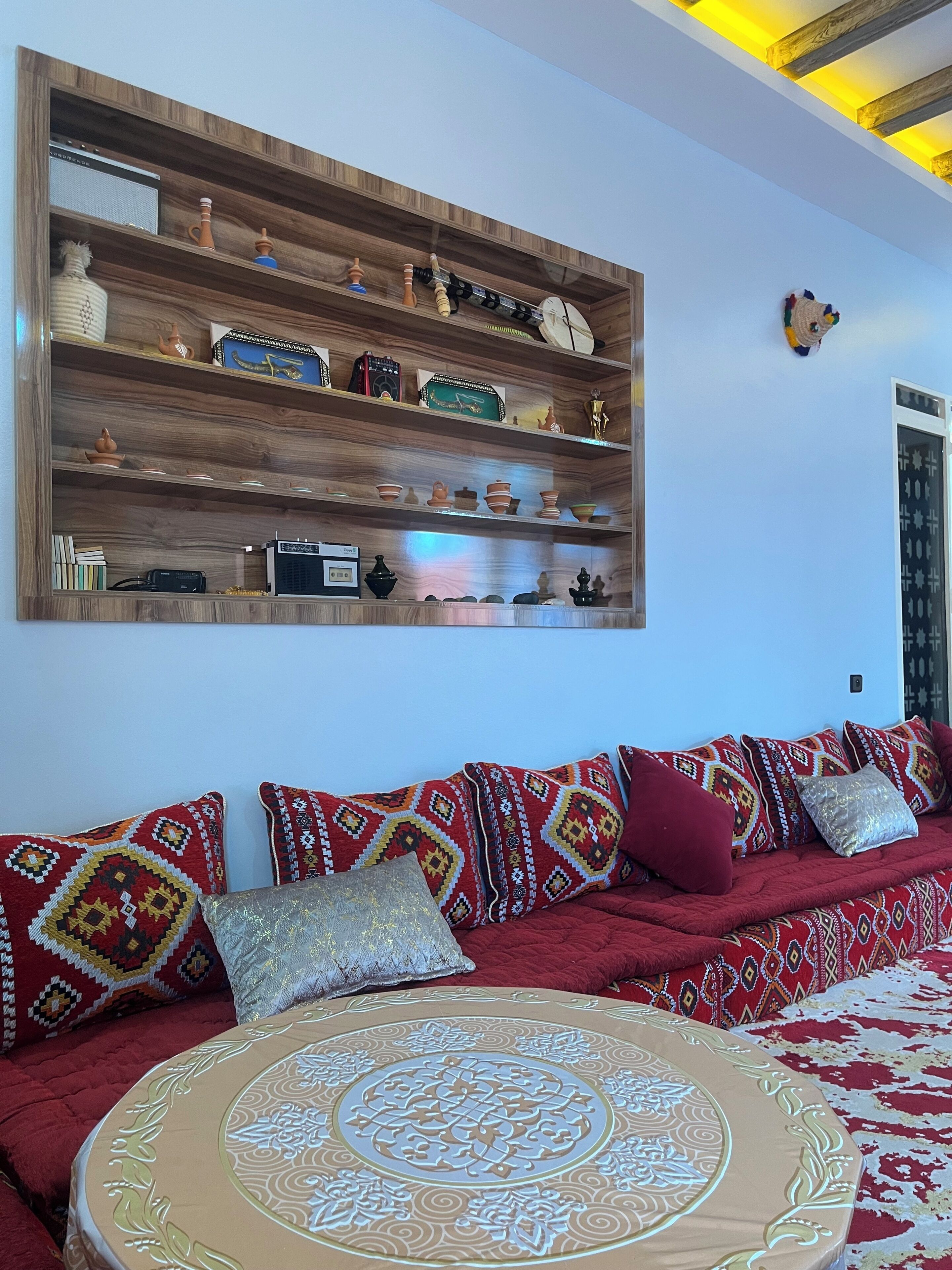 A room with a wooden shelf displaying various items, a red sofa with patterned cushions, and a round table with a decorative cover.
