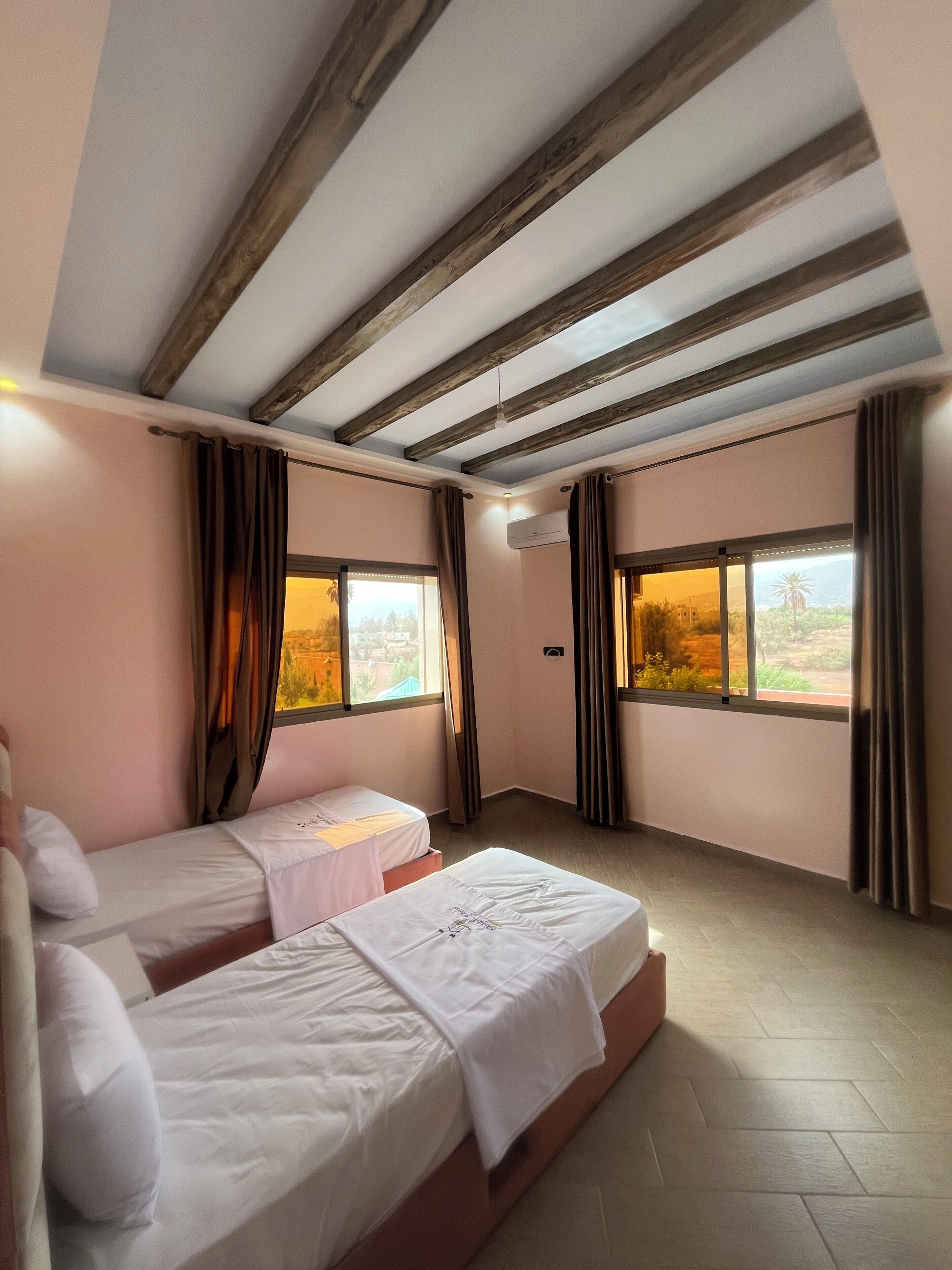 Una camera d'albergo con due letti singoli, un soffitto a vista con travi e ampie finestre con tende.