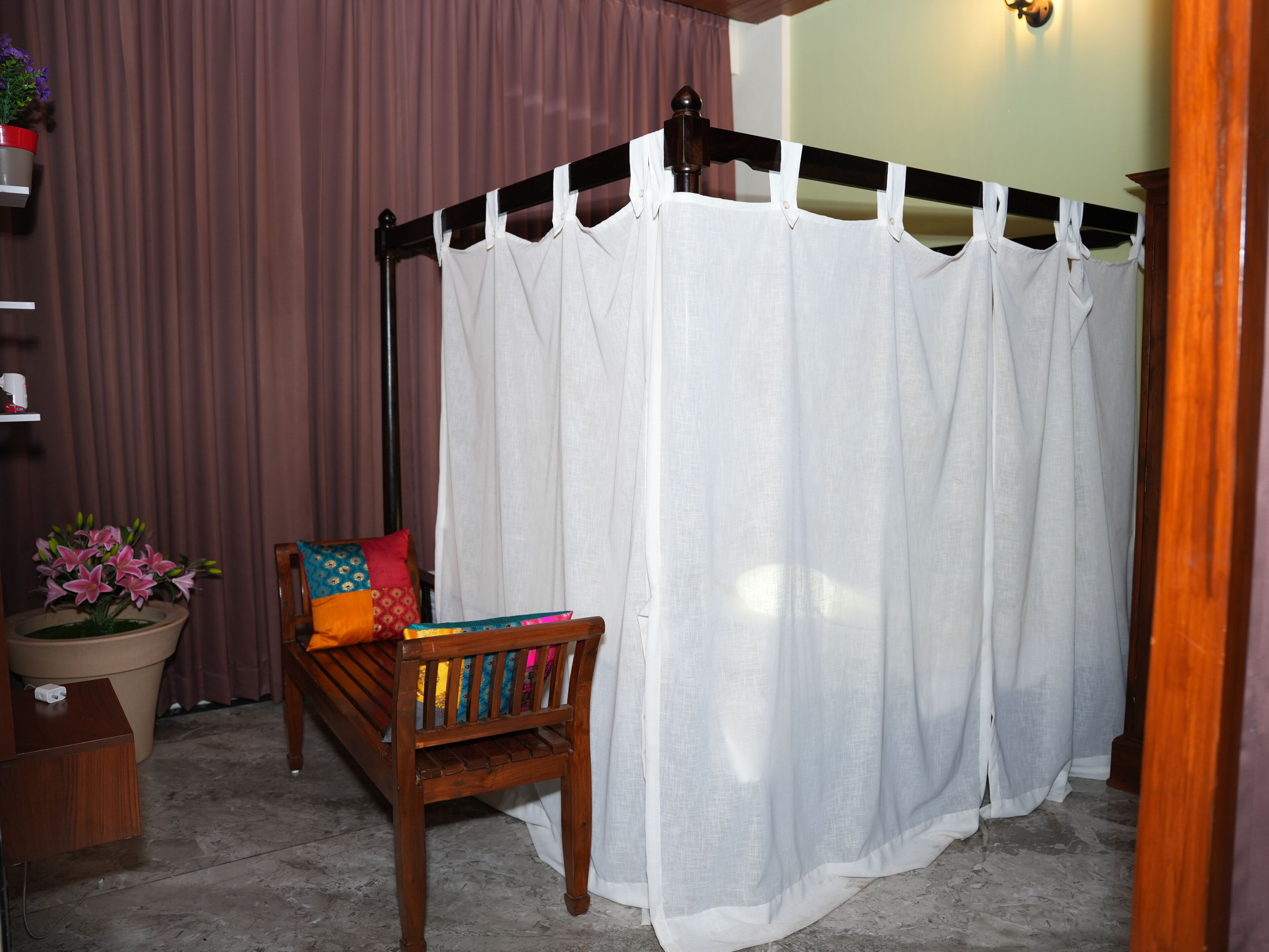 Um quarto com um banco de madeira, uma cama com dossel branca, um vaso de planta e uma prateleira fixada na parede.
