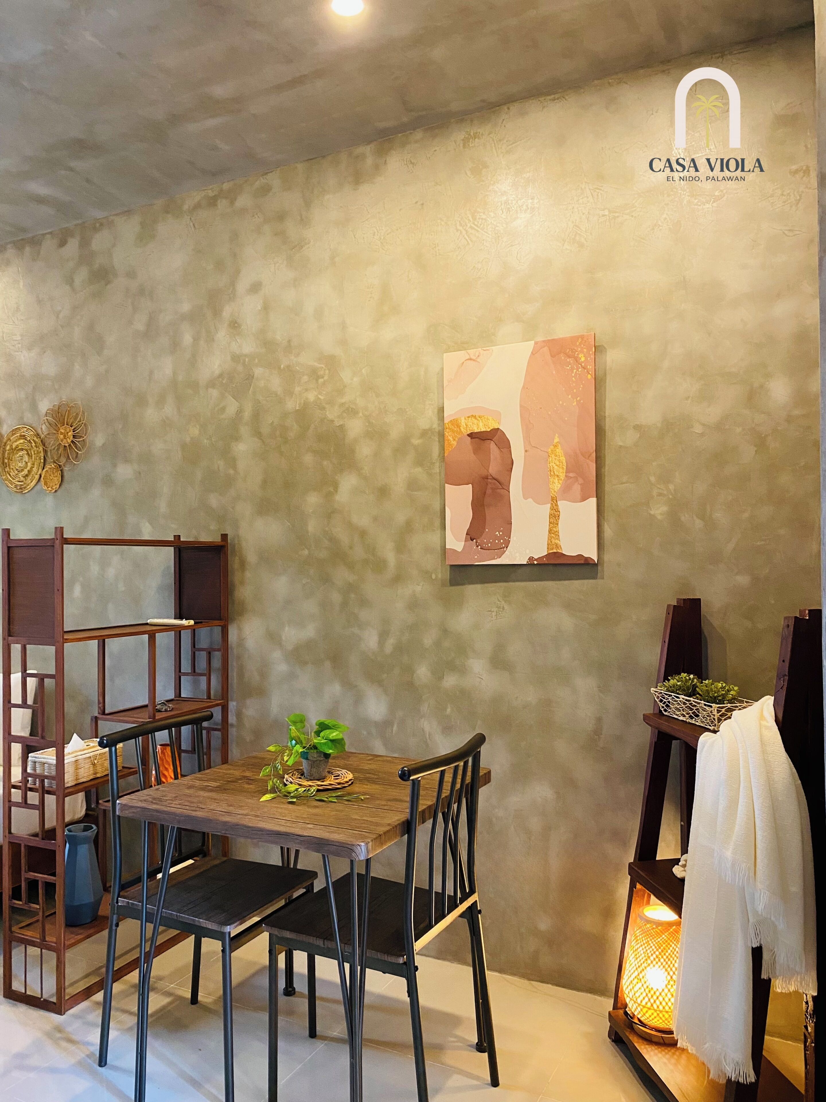 A dining area with a wooden table and chairs, a wall-mounted painting, and a shelf with decorative items.