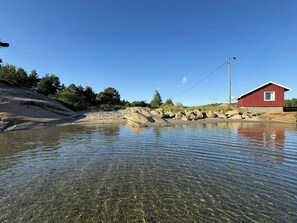 Ein klarer Fluss mit steiniger Uferlinie, ein rotes Gebäude und Bäume im Hintergrund.