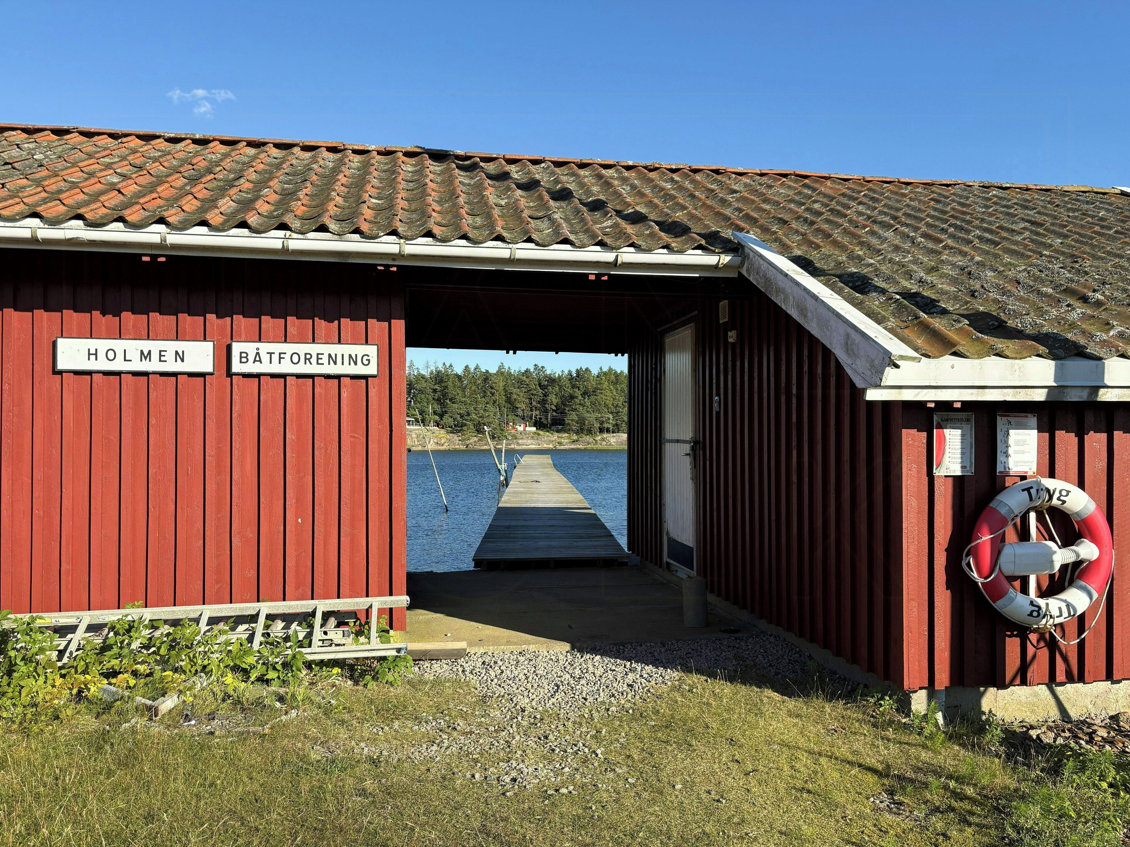 Ein rotes Gebäude mit den Schildern 'HOLMEN' und 'BÄTFORENING', eine anlegende Mole und ein Rettungsring auf der rechten Seite.