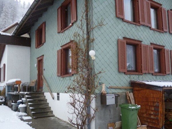 A multi-story building with a green facade and brown shutters, a staircase leading to the entrance, and a green trash bin. - Privatzimmer Hobi, (Valens), by Interhome (Valens)