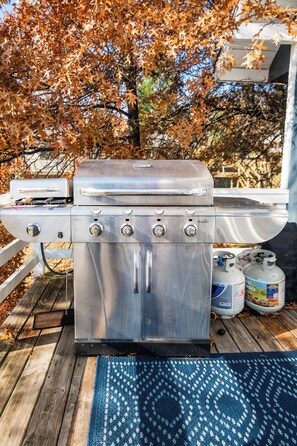 A stainless steel barbecue grill with four burners, two propane tanks, and a blue patterned rug on a wooden deck. - Charming 4-bedroom house with AC in Boyne Falls (Boyne Falls)