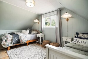 Um quarto com teto inclinado, uma cama com roupa de cama florida, uma mesinha de cabeceira e uma janela com cortinas.