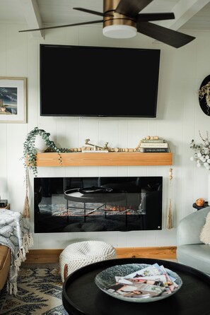 A living room with a flat-screen TV mounted above a fireplace, a ceiling fan, and a bookshelf with books. - Charming 4-bedroom house with AC in Boyne Falls (Boyne Falls)