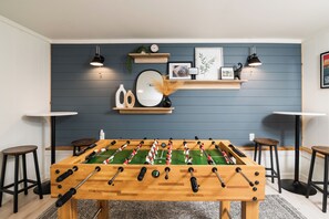 Quarto com mesa de futebol, dois banquetes, uma mesinha e uma prateleira fixada na parede com objetos decorativos.