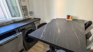 Ett kompakt kök med tvättmaskin, ett matbord med en bordsskiva i marmor och ett fönster med gardiner.