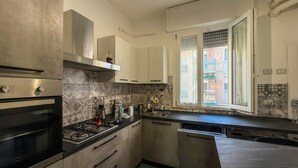 Ett modernt kök med gasspis, ugn och diskmaskin, ett fönster med utsikt över en byggnad och en kakelbeklädd vägg bakom arbetsbänken.