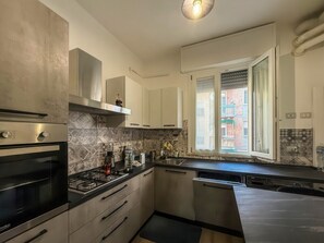 A modern kitchen with a gas stove, oven, and dishwasher, a window with a view of a building, and a tiled backsplash. - Experience Ferrara - Elegant and Bright Apartment Just a Stone's Throw from Downtown (Ferrara)