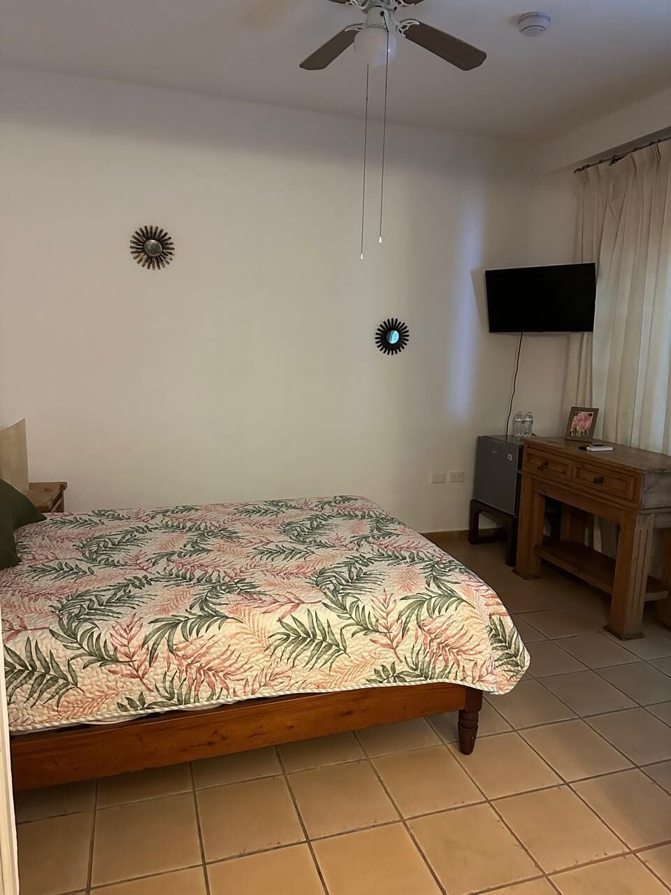 Une chambre à coucher avec un lit, un ventilateur de plafond, une télévision, une petite armoire et une fenêtre avec des rideaux.