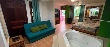 A room with a bed, a sofa, a small table, a bathtub, and a door leading to another room.