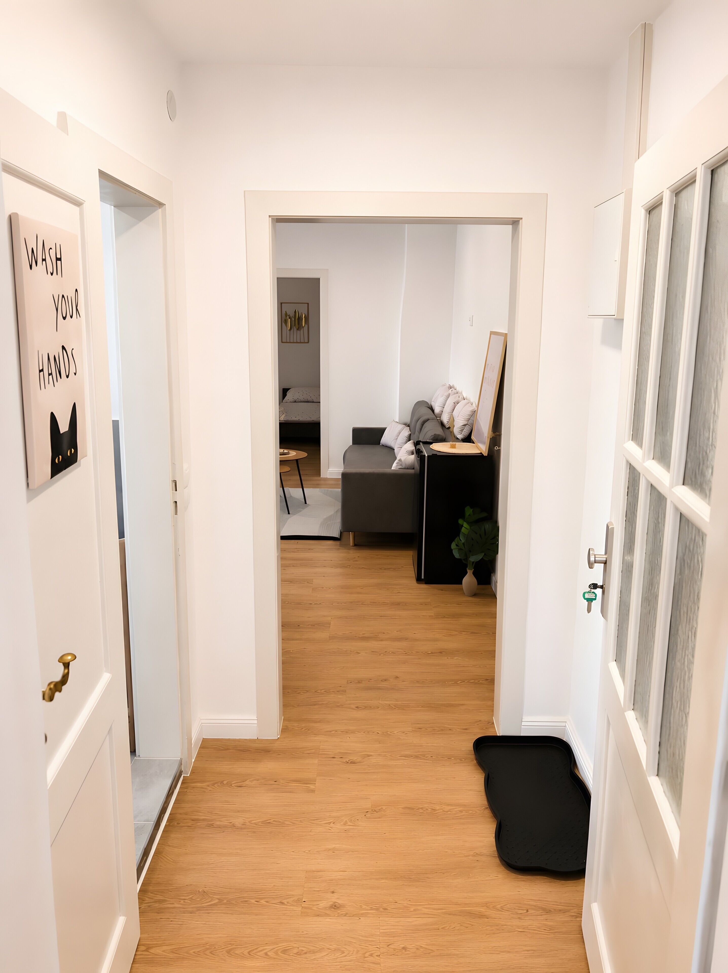 Ein Flur mit Holzboden, einer schwarzen Matte am Eingang, einem Schild an der linken Tür und einem Bett im Hintergrund.