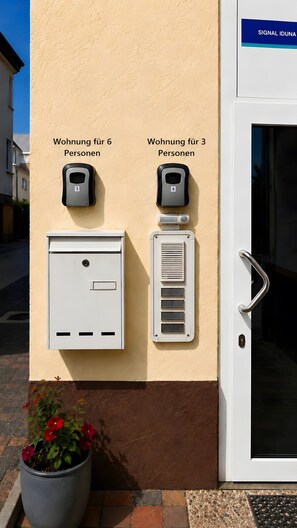 Entrada de un edificio con una puerta, un buzón y dos dispositivos en la pared indicando la capacidad de distintas habitaciones.