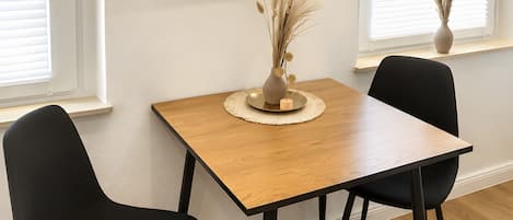Une pièce avec un téléviseur à écran plat fixé au mur, une table en bois avec deux chaises noires et un vase contenant des fleurs séchées, posé sur un rebord de fenêtre.