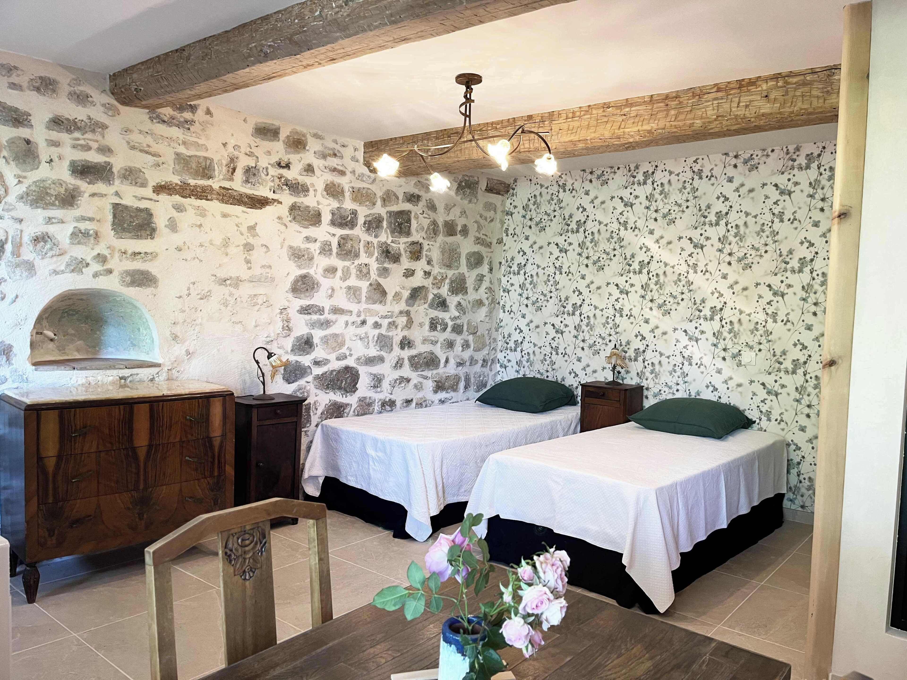 A rustic room with stone walls, a wooden ceiling, a bed, a dresser, and a table with flowers.
