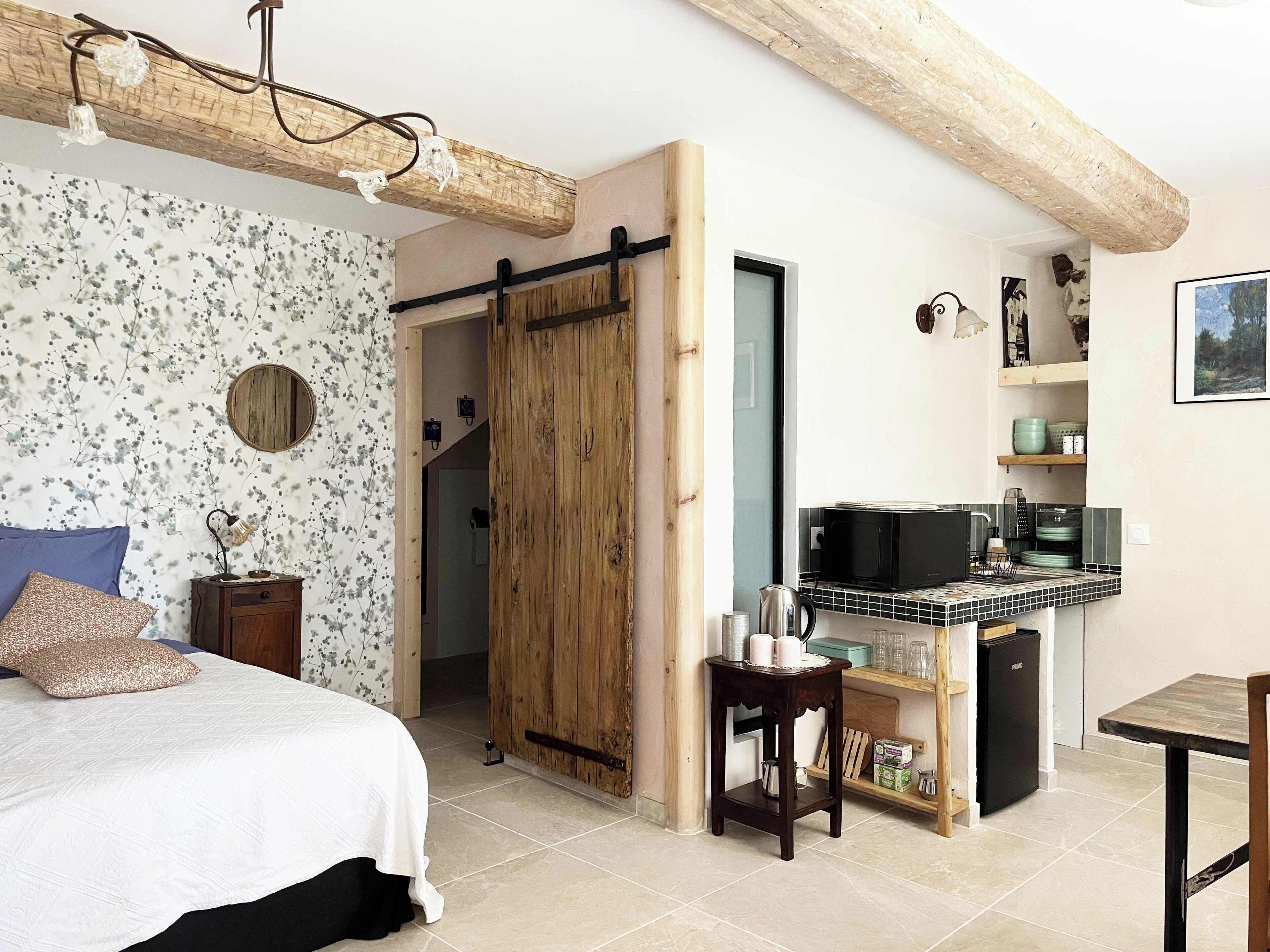 A bedroom with a bed, a bedside table, a wooden wardrobe, a kitchen area with a microwave, and a dining table.