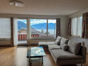 Ein Wohnzimmer mit Glastisch, grauem Sofa und Blick auf Berge und See.
