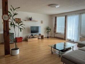 Ein Wohnzimmer mit Holzboden, einem Glastisch, einer Couch, einem Fernseher und Topfpflanzen.