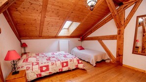 Un dormitorio con techo inclinado, vigas de madera, una cama con colcha, una mesita de noche con lámpara y una ventana.