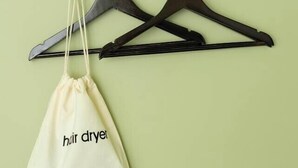 Eine an der Wand befestigte Holzablage mit drei schwarzen Kleiderbügeln und einer weißen Baumwolltasche mit dem Aufdruck 'hair dryer'.