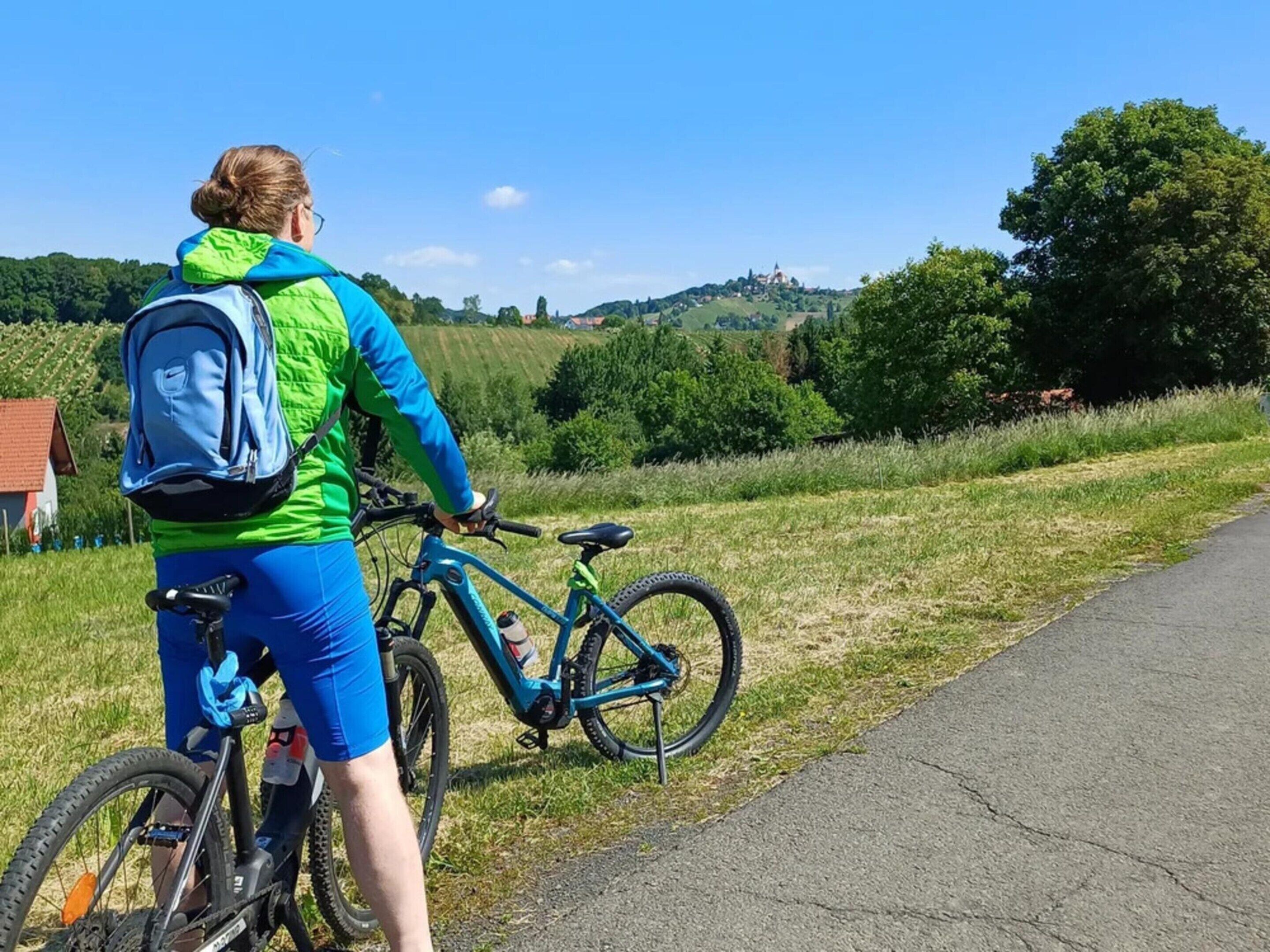 Une personne à vélo, portant un sac à dos, se trouve sur un chemin pavé bordé de gazon, sous un ciel dégagé, avec un paysage vallonné en arrière-plan.