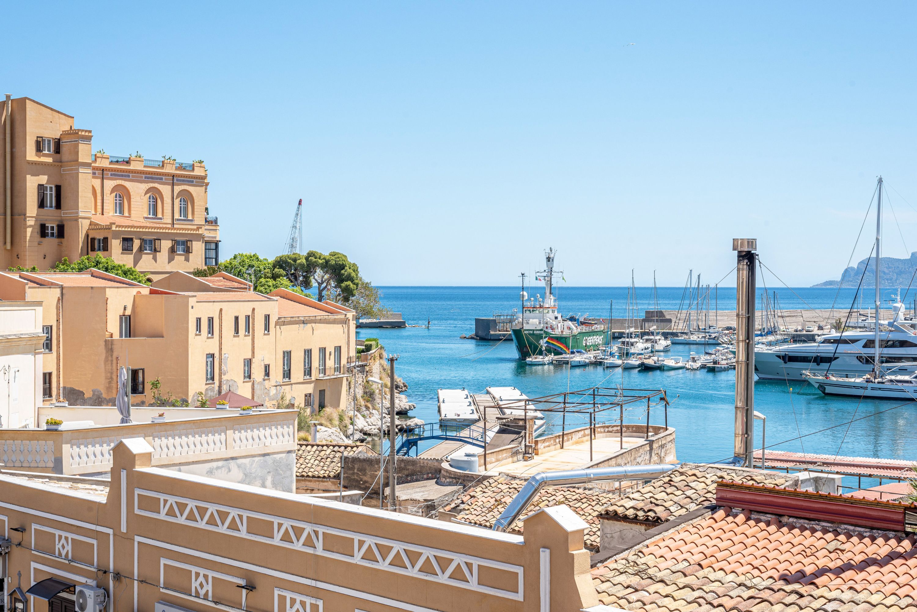 Una marina costera con barcos amarrados, un edificio grande con ventanas arqueadas y un cielo azul despejado.