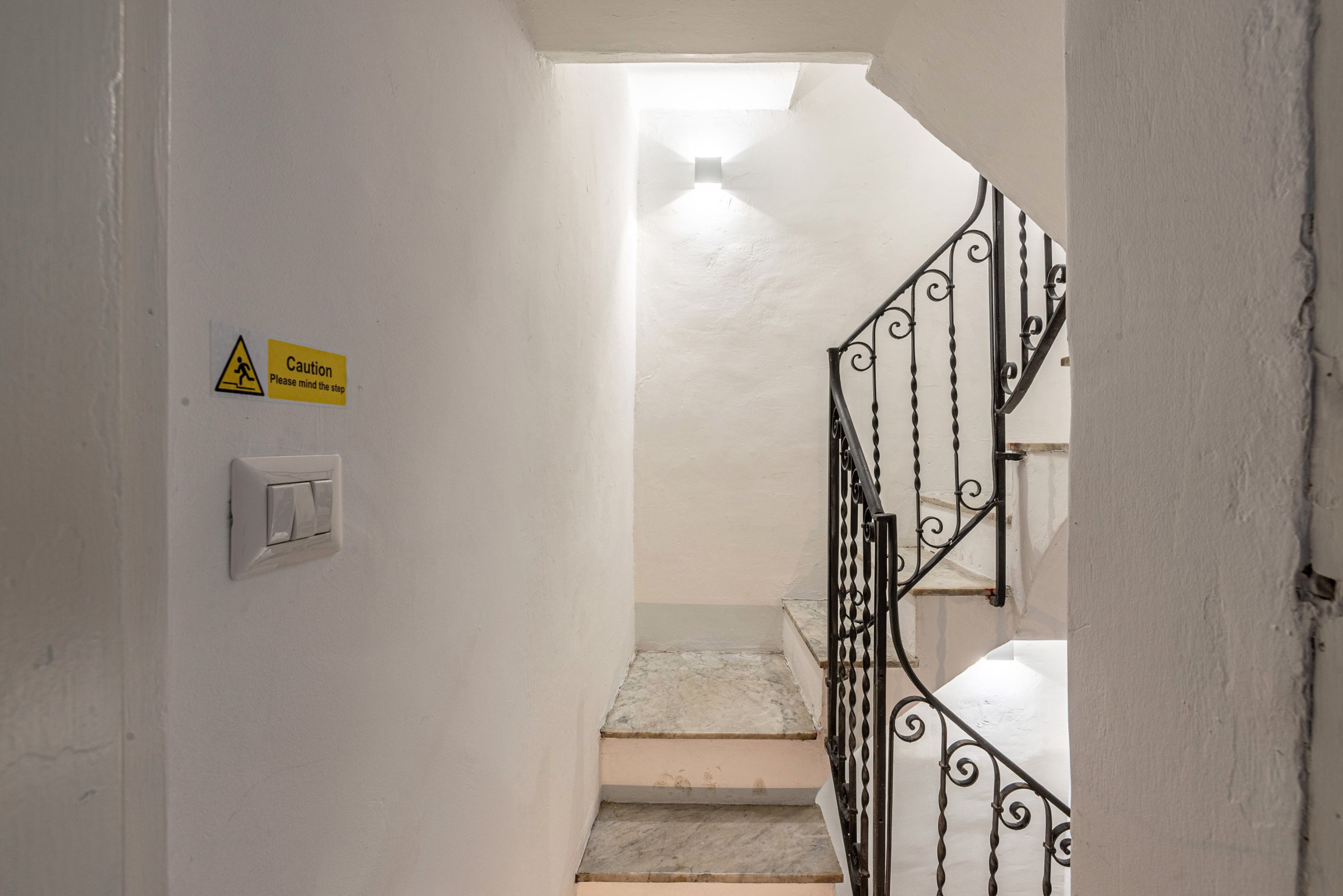 Una escalera con un cartel de precaución, un interruptor de luz y una barandilla metálica.