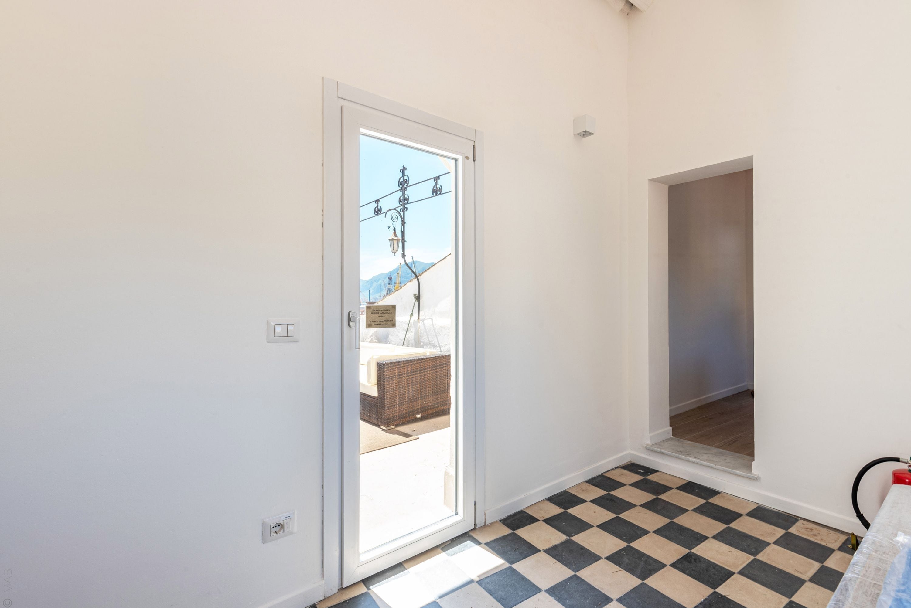Una habitación con piso a cuadros, una puerta blanca y vistas a un balcón con una calle y montañas.