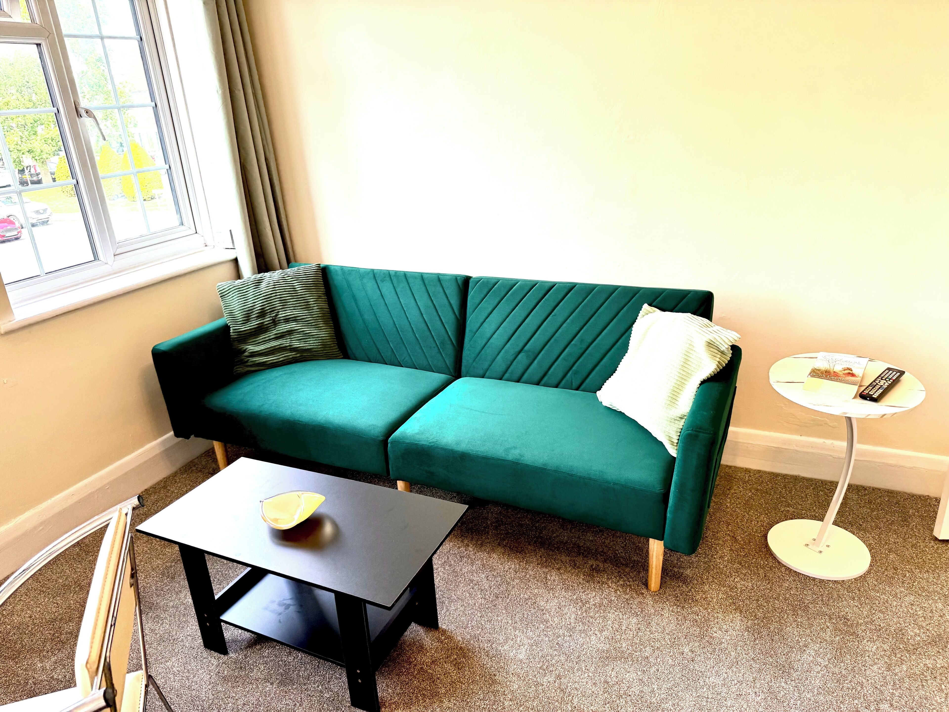 A teal sofa with a striped pillow, a small table with a bowl, and a round side table with a remote control.