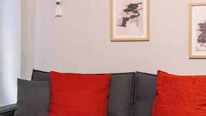 Uma sala de estar moderna com um sofá cinzento, almofadas vermelhas, uma mesa de centro de madeira com croissants e sumo de laranja, e um aparelho de ar condicionado na parede.