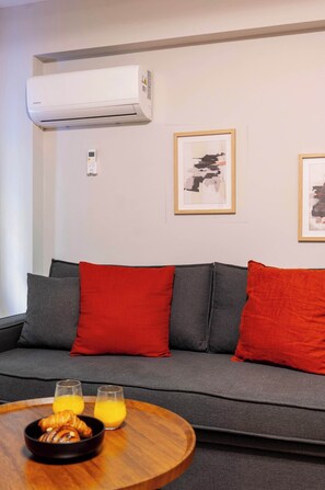 Ein modernes Wohnzimmer mit einem grauen Sofa, roten Kissen, einem Holztisch mit Croissants und Orangensaft, und einer Klimaanlage an der Wand.