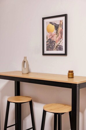 Ein schlichter Raum mit einem Holztisch und zwei Hocker, einem gerahmten Bild an der Wand und einer kleinen Vase auf dem Tisch.