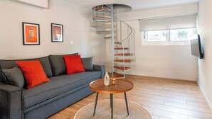 Una sala de estar moderna con un sofá gris, cojines rojos, una mesa de centro de madera, una escalera en espiral y obras de arte enmarcadas en la pared.