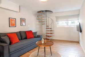 Ein modernes Wohnzimmer mit grauem Sofa, roten Kissen, einem Holztisch und gerahmten Kunstwerken an der Wand, sowie einer Wendeltreppe.