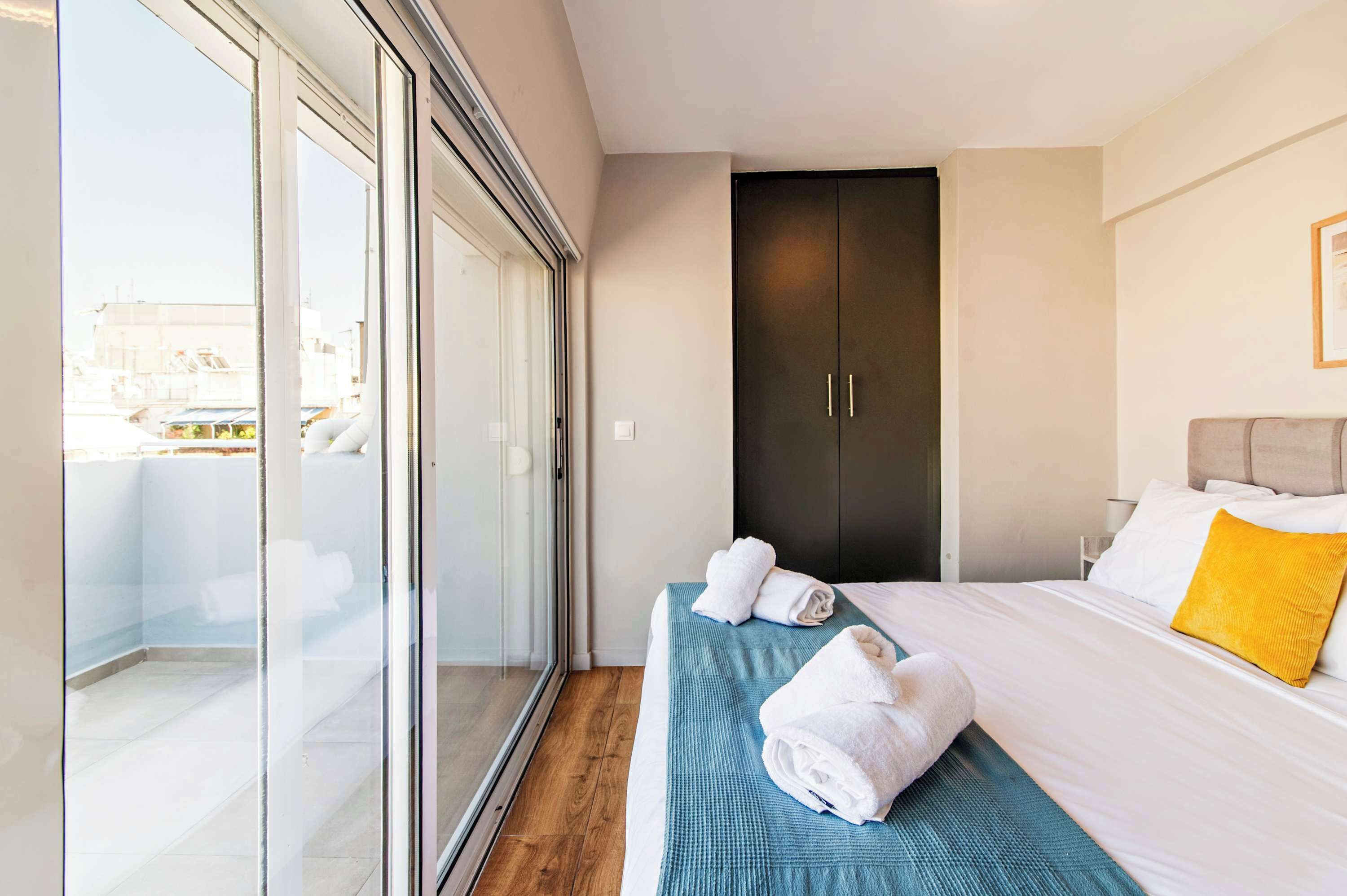 A modern hotel room with a large sliding glass door, a bed with white linens and a yellow pillow, and a dark-colored wardrobe.
