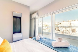 A hotel room with a large bed, a wardrobe, an air conditioning unit, and a view of the city through large windows. - Stylish Duplex Apartment Near Victoria Metro (Athens)