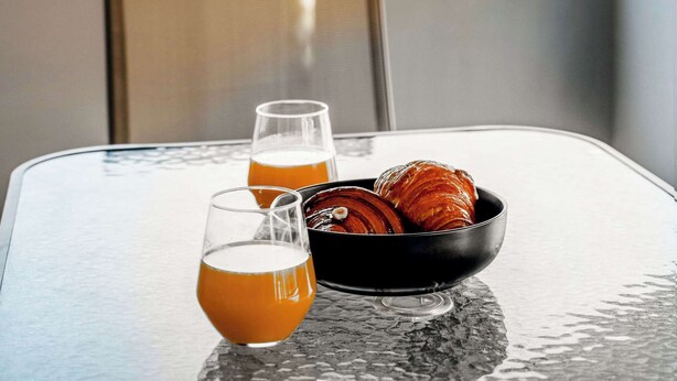 A table with a glass of orange juice, a glass of beer, and a bowl of croissants on a balcony overlooking a cityscape. - Modern Athens Apartment Near Attractions (Athens)