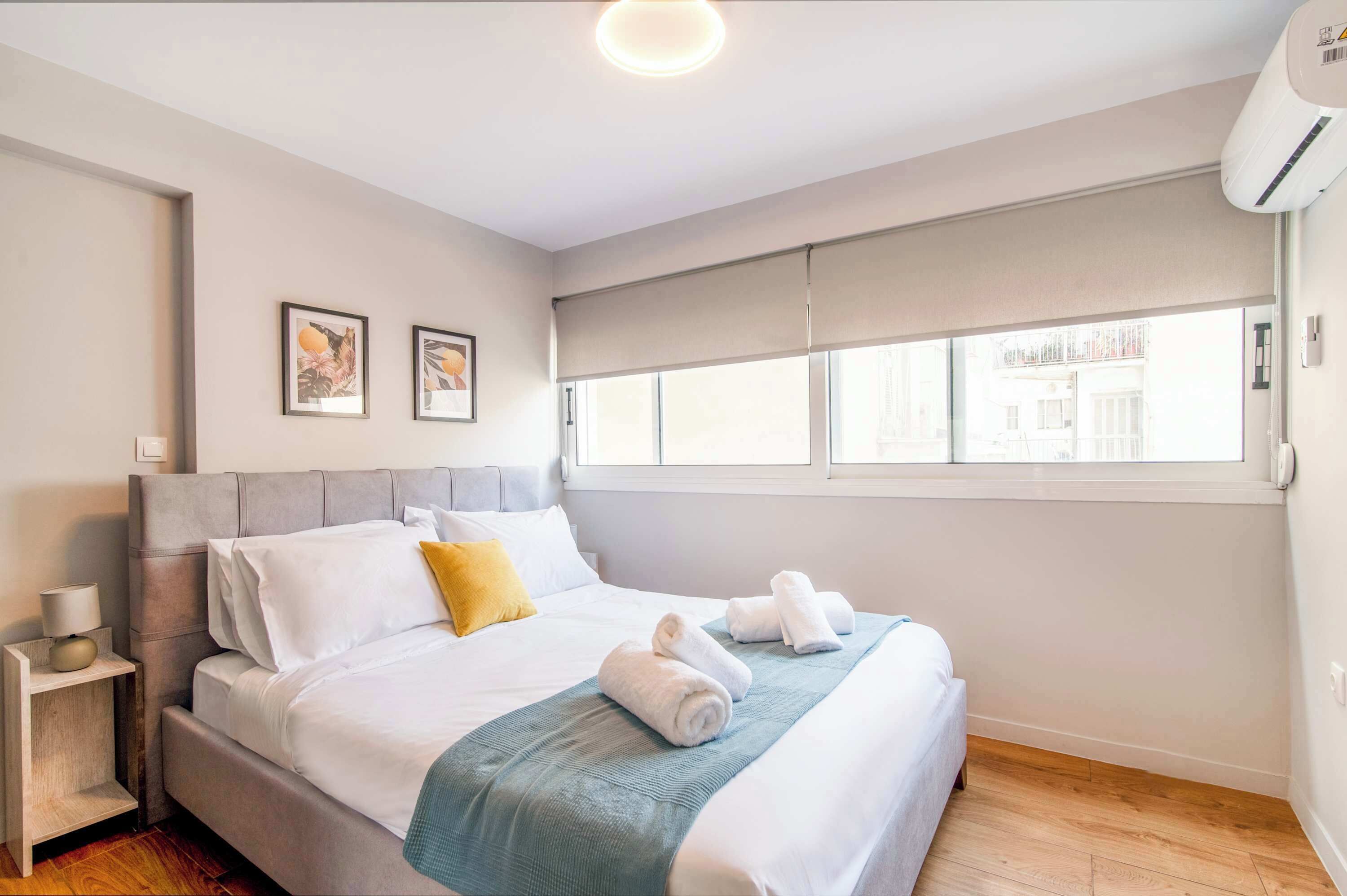 Une chambre moderne comprenant un lit, une table de chevet, une lampe, deux tableaux encadrés, une fenêtre avec vue et un appareil de climatisation.