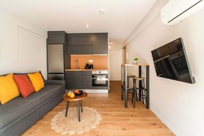 Een ruimtelijk ingerichte leefruimte met een bank, een klein rond tafeltje met drankjes, een kitchenette en een aan de muur bevestigde televisie.
