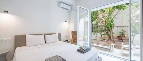 A modern hotel room with a large bed, a small bedside table, an air conditioning unit, and a sliding glass door leading to an outdoor area with plants and seating.