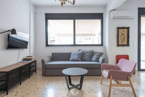 Ein modernes Wohnzimmer mit einer Couch, einem kleinen runden Tisch, einem rosafarbenen Sessel, einem an der Wand befestigten Fernseher und einem großen Fenster.