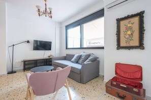 Ein Wohnzimmer mit grauem Sofa, einem rosafarbenen Sessel, einem Fernseher, einem Fenster und einem roten Koffer.