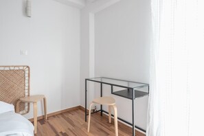 Ein Zimmer mit einem Bett, einem Glastisch, zwei hölzernen Hocker, einem Weidenkopfteil und einer weißen Wand.